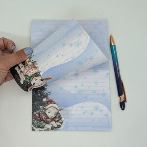 May include: A notepad with a winter theme, featuring a sleeping lamb illustration on the bottom and a similar lamb on the top corner. The notepad has blue and white snowflake designs and lined pages. A blue and gold pen is also present.