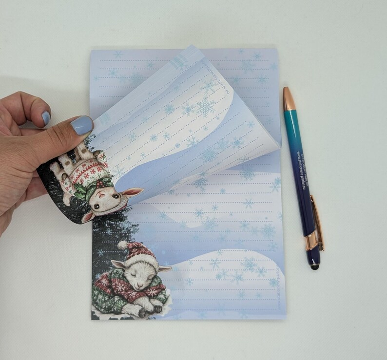 May include: A notepad with a winter theme, featuring a sleeping lamb illustration on the bottom and a similar lamb on the top corner. The notepad has blue and white snowflake designs and lined pages. A blue and gold pen is also present.