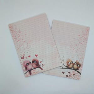 Pode incluir: Dois blocos de notas pautados com fundo rosa e detalhes de corações. Cada bloco de notas apresenta uma ilustração diferente de pássaros de desenho animado num galho. Um mostra dois pássaros, o outro três. Perfeito para escrever notas.