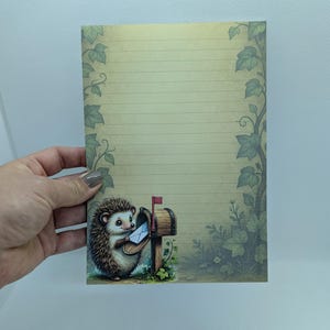May include: A lined stationery sheet with a whimsical illustration of a hedgehog delivering mail. The design features a brown mailbox, green foliage, and a light-colored background with decorative ivy. The stationery is held by a hand.