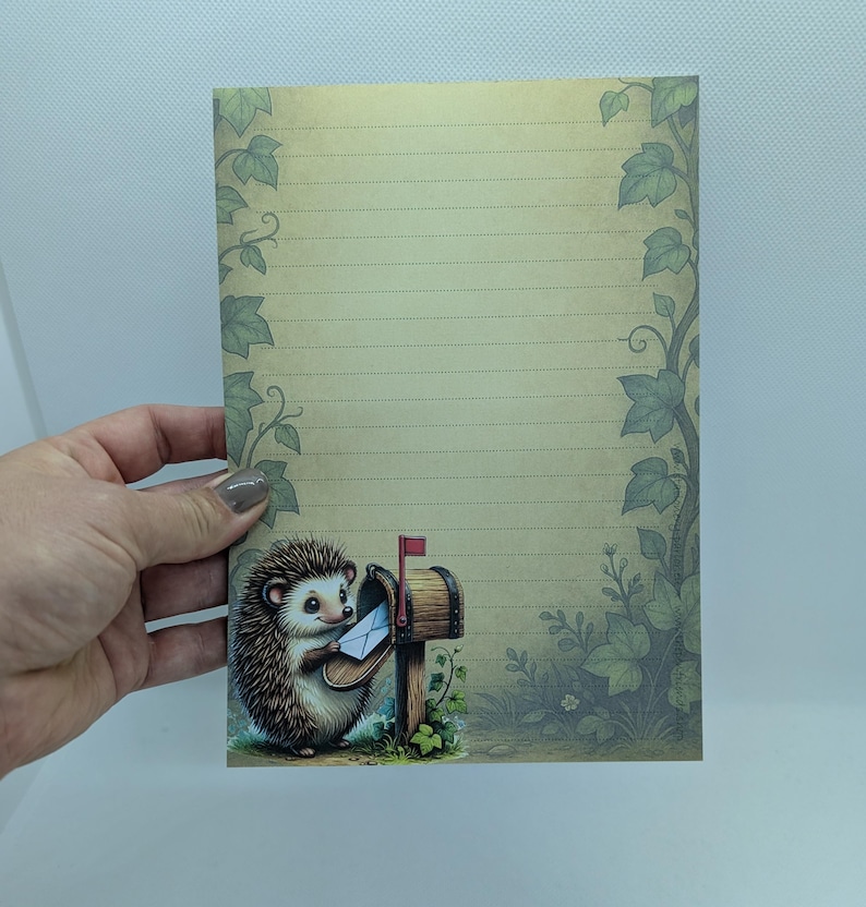 May include: A lined stationery sheet with a whimsical illustration of a hedgehog delivering mail. The design features a brown mailbox, green foliage, and a light-colored background with decorative ivy. The stationery is held by a hand.