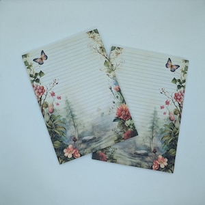 Pode incluir: Duas folhas de papel de carta pautado com um design floral e borboleta. A papelaria apresenta uma paisagem em aquarela com flores cor-de-rosa, folhagem verde e uma borboleta. O papel tem uma estética vintage.