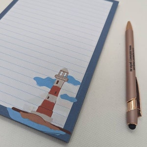 May include: A blue lined notepad with a lighthouse illustration and a white background. The notepad has a blue border and a white pen with a gold accent is next to it.