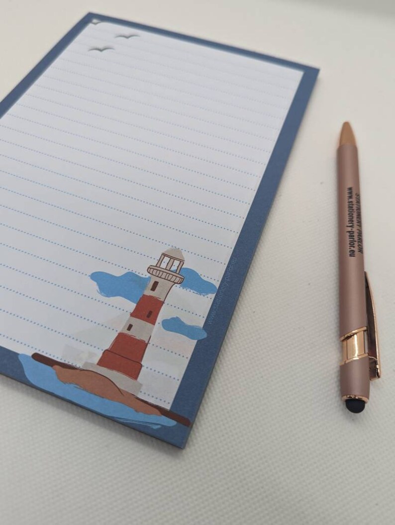 May include: A blue lined notepad with a lighthouse illustration and a white background. The notepad has a blue border and a white pen with a gold accent is next to it.
