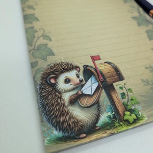 May include: A lined notepad with a whimsical illustration of a hedgehog delivering mail. The hedgehog is depicted with brown and tan quills, holding a letter at a wooden mailbox. The notepad has a vintage, rustic aesthetic with green leaf accents.