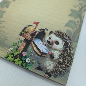 May include: A lined stationery sheet featuring a whimsical illustration of a hedgehog retrieving mail from a wooden mailbox. The artwork includes a red flag, flowers, and green foliage. The paper has a light tan background with a decorative border.