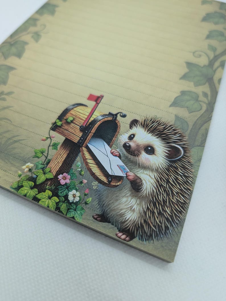 May include: A lined stationery sheet featuring a whimsical illustration of a hedgehog retrieving mail from a wooden mailbox. The artwork includes a red flag, flowers, and green foliage. The paper has a light tan background with a decorative border.