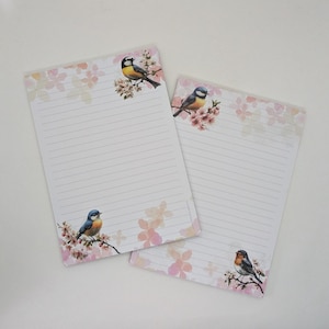 May include: Two lined notepads with a floral and bird design. The notepads have a white background with pink floral accents and illustrations of birds perched on branches. The birds are blue, yellow, and orange.