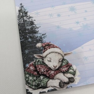 May include: A stationery sheet featuring a sleeping lamb wearing a red and white Santa hat and a green and red sweater. The background shows a snowy landscape with a tall evergreen tree.