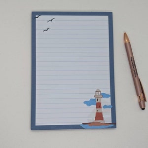 May include: A blue lined notepad with a lighthouse and seagulls illustration. The notepad has a blue border and a white interior. A gold pen is lying next to the notepad.