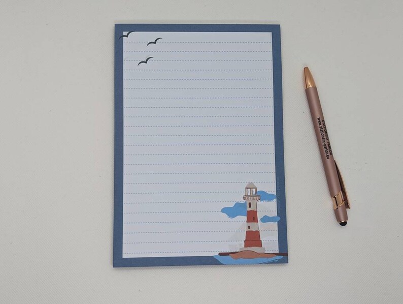 May include: A blue lined notepad with a lighthouse and seagulls illustration. The notepad has a blue border and a white interior. A gold pen is lying next to the notepad.