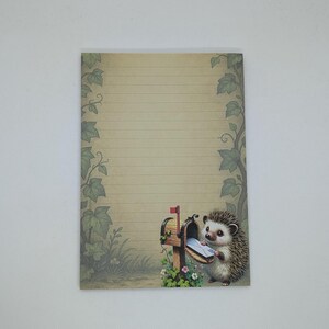 May include: A lined stationery sheet with a whimsical illustration of a hedgehog retrieving mail from a wooden mailbox. The mailbox has a red flag and is surrounded by flowers and greenery. The paper has a vintage, aged appearance.