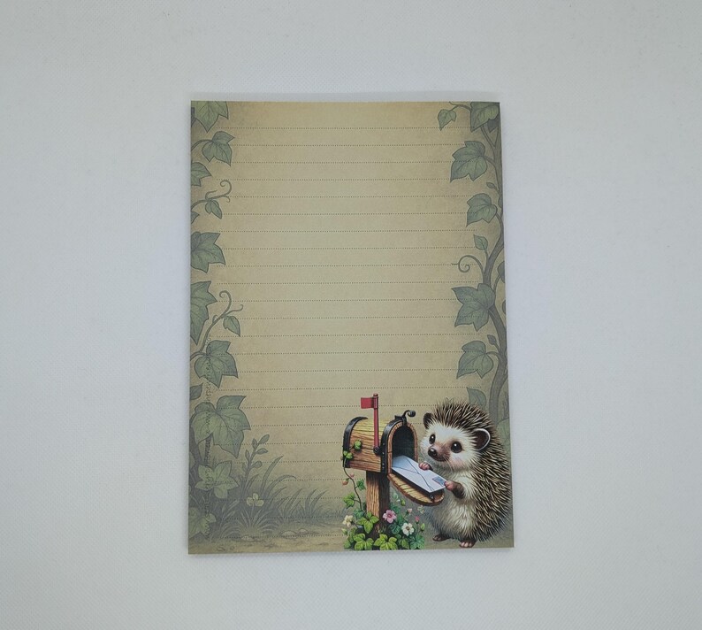 May include: A lined stationery sheet with a whimsical illustration of a hedgehog retrieving mail from a wooden mailbox. The mailbox has a red flag and is surrounded by flowers and greenery. The paper has a vintage, aged appearance.