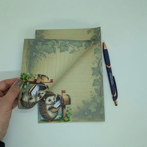 May include: Stationery set featuring a whimsical design with hedgehogs delivering mail. The set includes lined paper with a decorative border of leaves and a matching pen. The illustration has a vintage aesthetic with soft colors.