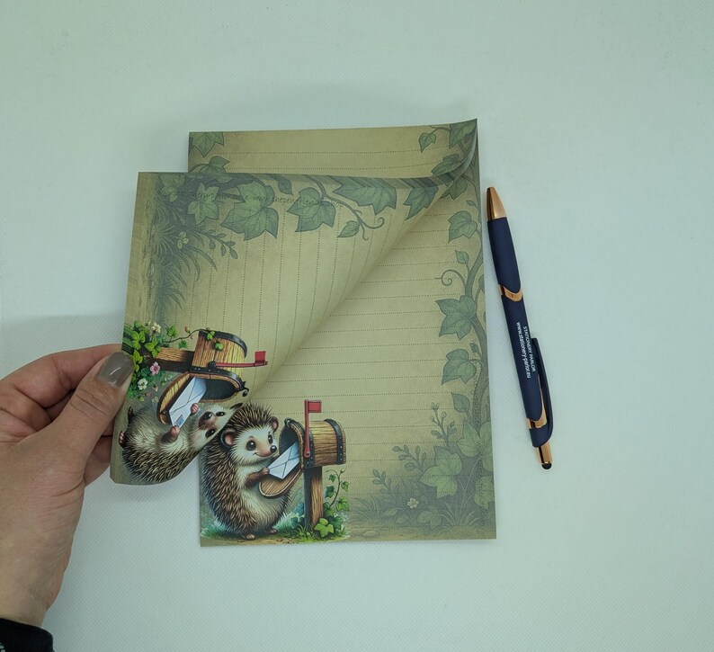 May include: Stationery set featuring a whimsical design with hedgehogs delivering mail. The set includes lined paper with a decorative border of leaves and a matching pen. The illustration has a vintage aesthetic with soft colors.