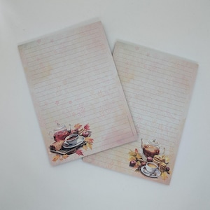 May include: Two lined notepads with a vintage design. The pads feature a watercolour illustration of a teapot, teacup, and autumn leaves. The paper has a cream colour with a pink floral pattern.