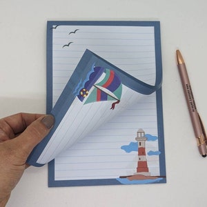 May include: A blue lined notepad with a sailboat and lighthouse illustration. The notepad has a blue border and a white background. The sailboat is colourful and has a red, white, and blue sail. The lighthouse is red and white with a black roof. The notepad is folded in half and the top half is flipped over to reveal the lined paper.