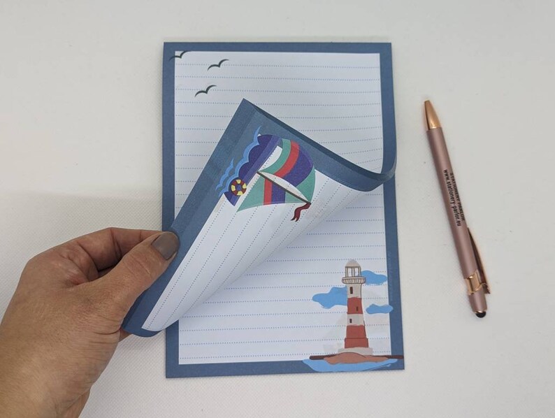 May include: A blue lined notepad with a sailboat and lighthouse illustration. The notepad has a blue border and a white background. The sailboat is colourful and has a red, white, and blue sail. The lighthouse is red and white with a black roof. The notepad is folded in half and the top half is flipped over to reveal the lined paper.