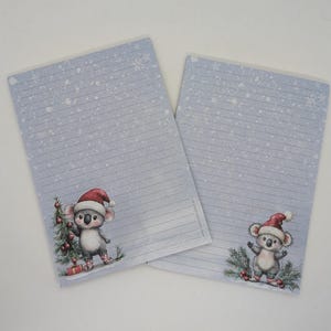 Op de afbeelding: Twee gelijnde notitieblokken met een lichtblauwe achtergrond en een kerstthema. Elk notitieblok heeft een cartoon koala met een kerstmuts en een kerstboom. Sneeuwvlokken zijn verspreid over het papier.