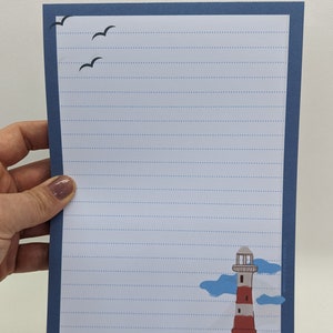 May include: Blue lined paper with a white border and a lighthouse illustration in the bottom right corner. The lighthouse is red and white with a black roof and a blue sky with white clouds behind it. There are three black birds flying in the top left corner.