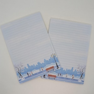 May include: Two lined notepads with a winter scene border. The border features a blue cityscape, people ice skating, benches, and a lamppost. The notepads have a light blue background with horizontal lines.
