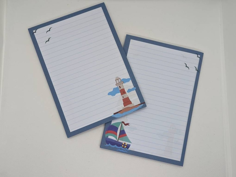 May include: Two blue lined notepads with a nautical theme. One notepad features a lighthouse with clouds and the other features a sailboat with blue, red and white sails.
