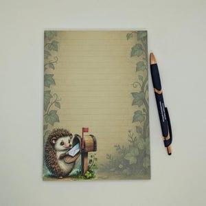 May include: A lined notepad with a whimsical illustration of a hedgehog posting mail. The notepad has a beige background with green ivy accents. A black and gold pen rests on the right side of the notepad.