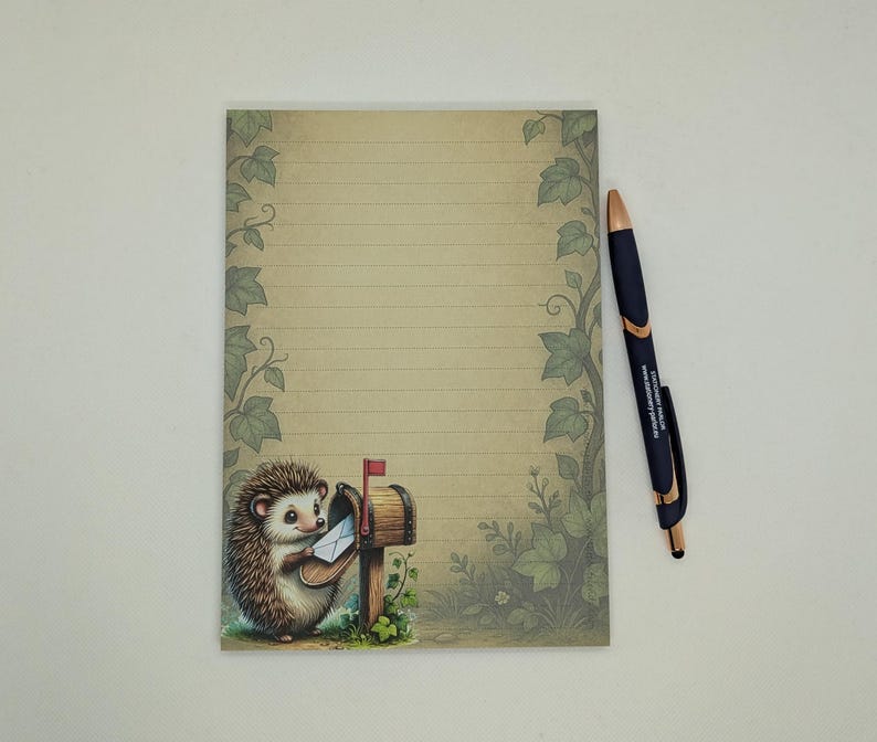 May include: A lined notepad with a whimsical illustration of a hedgehog posting mail. The notepad has a beige background with green ivy accents. A black and gold pen rests on the right side of the notepad.
