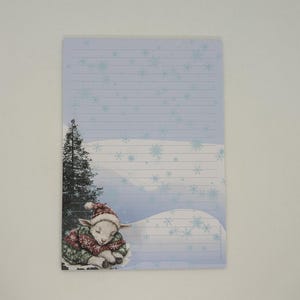 May include: A lined notepad with a winter theme. The illustration features a sleeping lamb wearing a red and green sweater and hat, with a snowy landscape and a tall evergreen tree. The background is light blue with snowflakes.