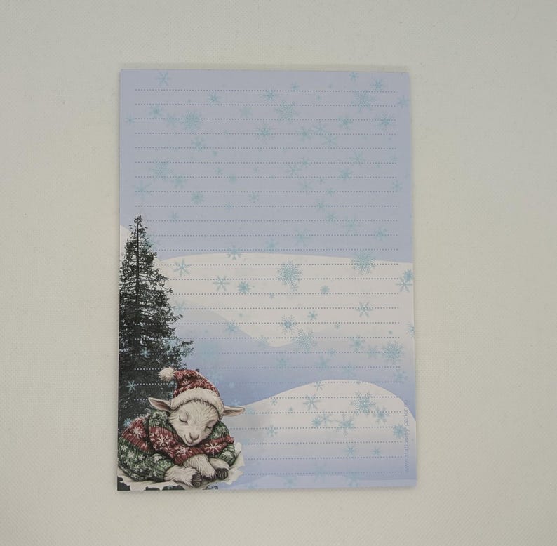 May include: A lined notepad with a winter theme. The illustration features a sleeping lamb wearing a red and green sweater and hat, with a snowy landscape and a tall evergreen tree. The background is light blue with snowflakes.