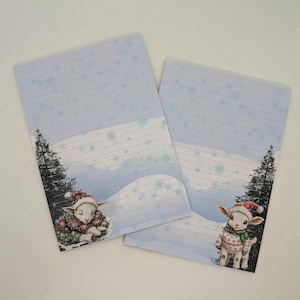 May include: Two lined stationery sheets with a winter theme. Each sheet features a light blue background with snowflakes and a cartoon lamb wearing a winter hat and sweater. A dark green pine tree is on the left side of each sheet.