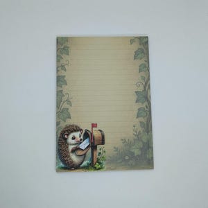 May include: A lined notepad with a whimsical illustration of a hedgehog posting a letter. The notepad features a vintage-style design with a beige background, green leaf borders, and a small mailbox.