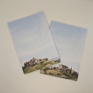 Op de afbeelding: Twee gelijnde notitieblokken met een schilderachtig landschapsontwerp. De onderkant van elk notitieblok heeft een aquarel schilderij van een dorp met wijngaarden en glooiende heuvels. De lucht is lichtblauw met stippellijnen om op te schrijven.