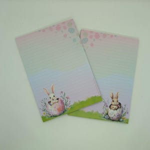 Op de afbeelding: Twee gelinieerde notitieblokken met een Paasthema. Elk notitieblok heeft een pastelkleurige gradiënt achtergrond, een konijn dat uit een ei komt en decoratieve paasei-ontwerpen. De notitieblokken hebben een groene grasrand.
