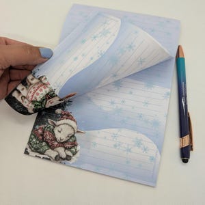 May include: A notepad with a winter theme, featuring a sleeping lamb illustration wearing a hat and sweater. The notepad has a light blue background with snowflakes and lined pages. A blue and gold pen is next to the notepad.