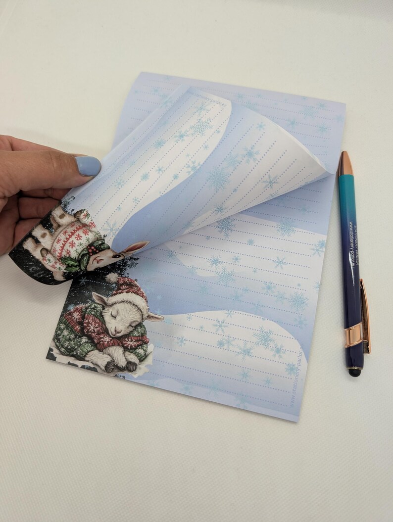 May include: A notepad with a winter theme, featuring a sleeping lamb illustration wearing a hat and sweater. The notepad has a light blue background with snowflakes and lined pages. A blue and gold pen is next to the notepad.