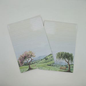 May include: Two lined stationery sheets with a landscape design. One sheet features a tree with pink blossoms and a swing, the other a weeping willow with a swing. The background shows green hills and a blue sky.