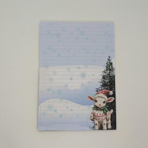 May include: A lined notepad with a winter scene featuring a lamb wearing a Santa hat and sweater. The notepad has a light blue background with snowflakes and a snowy landscape.