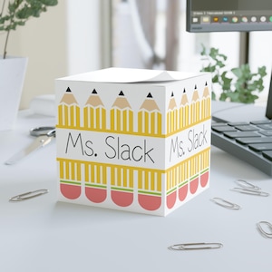 May include: A white notepad cube with a pencil design and the text "Ms. Slack" in black. The cube features yellow pencils with black tips and red erasers. The notepad is on a white desk with paper clips.