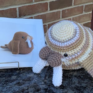 May include: A crocheted walrus plush toy featuring a striped pattern in shades of cream, tan, and brown. The plush has two dark eyes, a brown muzzle, and two long white tusks. It is positioned next to an open book displaying a photo of a walrus.