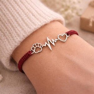 Op de afbeelding: Een bordeauxrood gevlochten armband met zilveren bedels. De bedels zijn onder meer een pootafdruk, een hartslaglijn en een hart. De armband wordt om een pols gedragen, met een crèmekleurige trui op de achtergrond. Een accessoire met een huisdierthema.