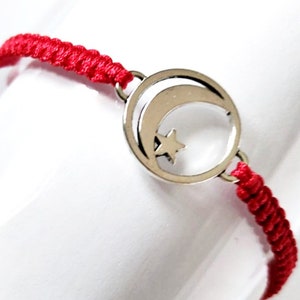 Op de afbeelding: Rood koord armband met een zilveren bedel in de vorm van een halve maan en een ster.