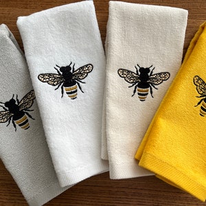 May include: Four embroidered hand towels in shades of gray, white, cream, and yellow. Each towel features a detailed black and gold bee design. The towels are made of a soft, absorbent material, perfect for the kitchen or bathroom.