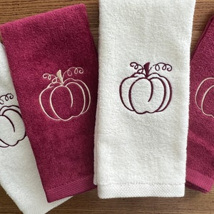 May include: Four folded hand towels, two white and two burgundy, with embroidered pumpkin designs. The pumpkins have a stem and curly vines. The towels are made of a soft, absorbent material, perfect for the fall season.
