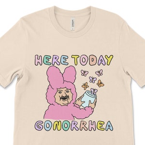 May include: A beige t-shirt with a pink bunny rabbit wearing a blue and white can. The bunny has a mustache and is holding the can up to its mouth. The text "HERE TODAY GONORRHEA" is printed on the shirt in rainbow colors.