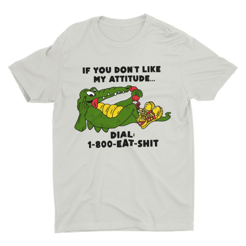 以下が含まれることがあります： 薄いグレーのTシャツに、赤い電話を持った緑色のワニの漫画が描かれています。ワニは赤い靴を履いています。Tシャツには「If you don't like my attitude... Dial: 1-800-EAT-SHIT」という文字が書かれています。