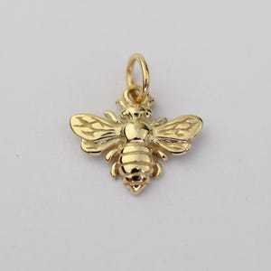 May include: A polished gold bee charm featuring detailed wing textures and a segmented body. The charm includes a small circular jump ring at the top for attachment, showcasing a shiny metallic finish and a classic insect shape design.
