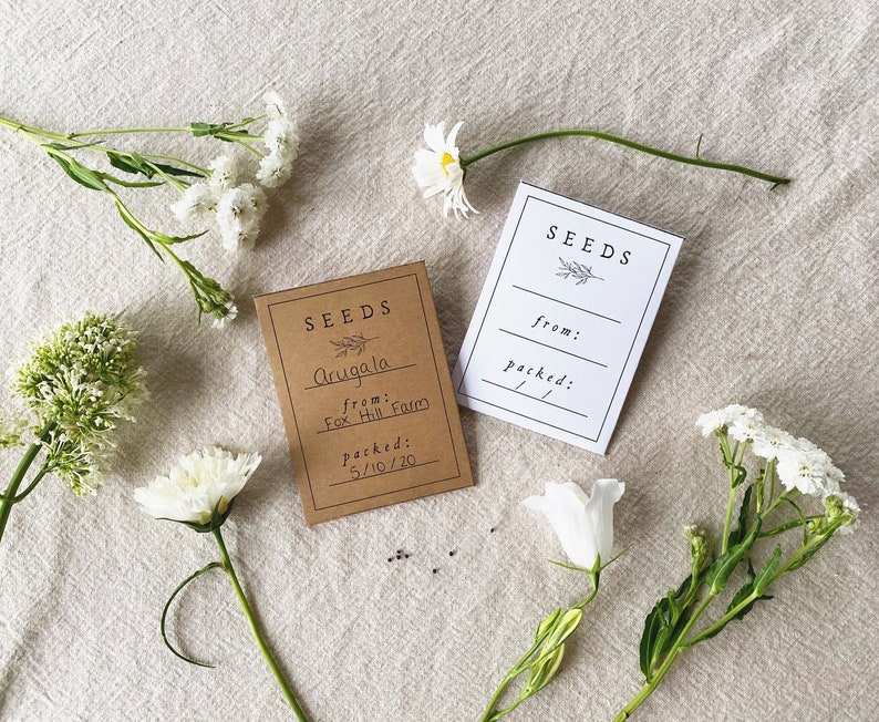 May include: Two seed packets, one brown and one white, with floral accents and the word "SEEDS". The brown packet reads "Arugala from Fox Hill Farm, packed 5/10/20". White flowers and greenery are arranged around the packets on a neutral linen surface.