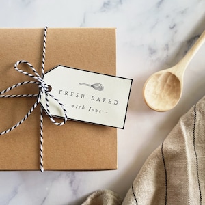 以下が含まれることがあります： 黒と白の紐で結ばれた茶色の段ボール箱。タグには「FRESH BAKED with love」と書かれています。木のスプーンとストライプのリネン布も写っています。