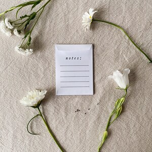 May include: A white envelope with the word "notes:" printed on it, with lines for writing. The envelope is surrounded by white flowers and green stems, arranged on a beige linen surface.